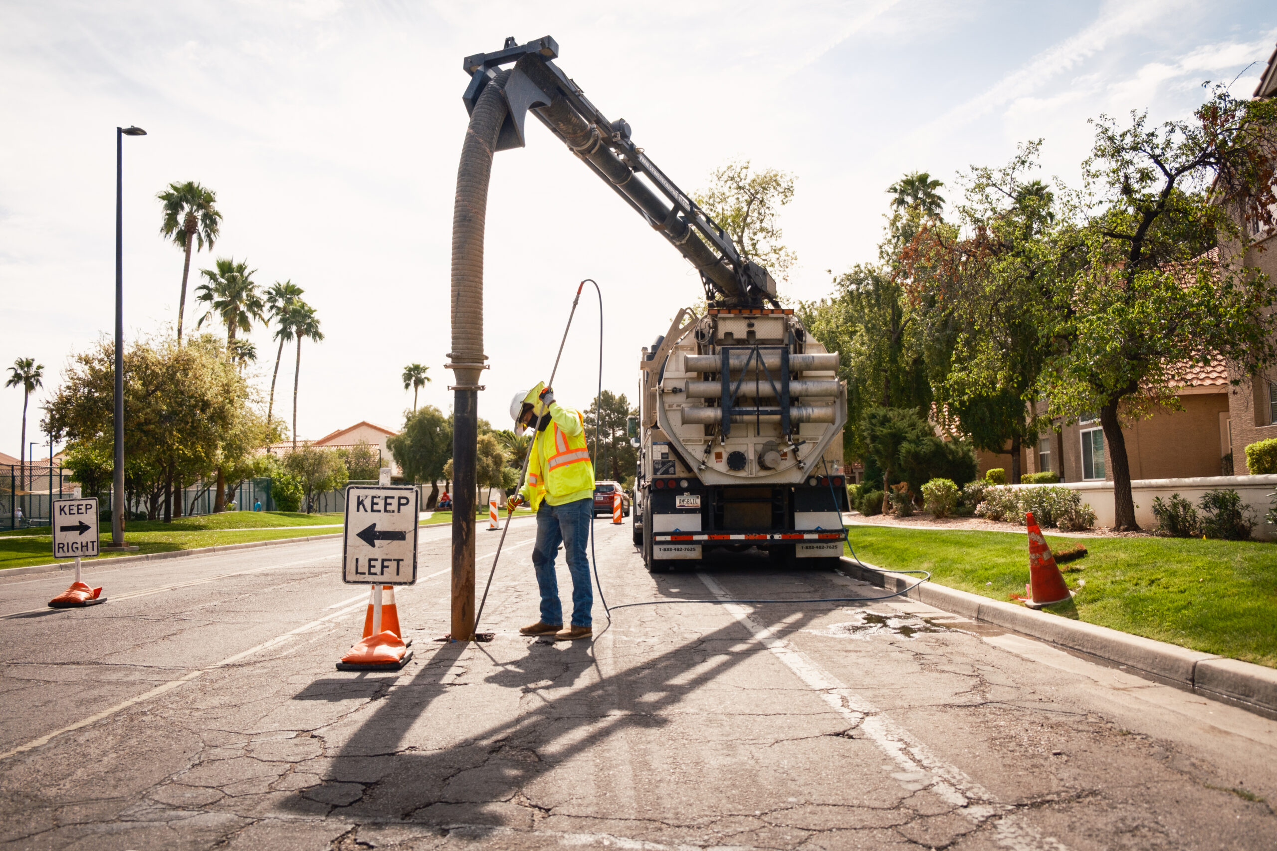 A construction worker in a high-visibility vest and hard hat operates a large vacuum truck, holding a hose into a hole in the road, with traffic cones and directional signs guiding vehicles around the work zone.