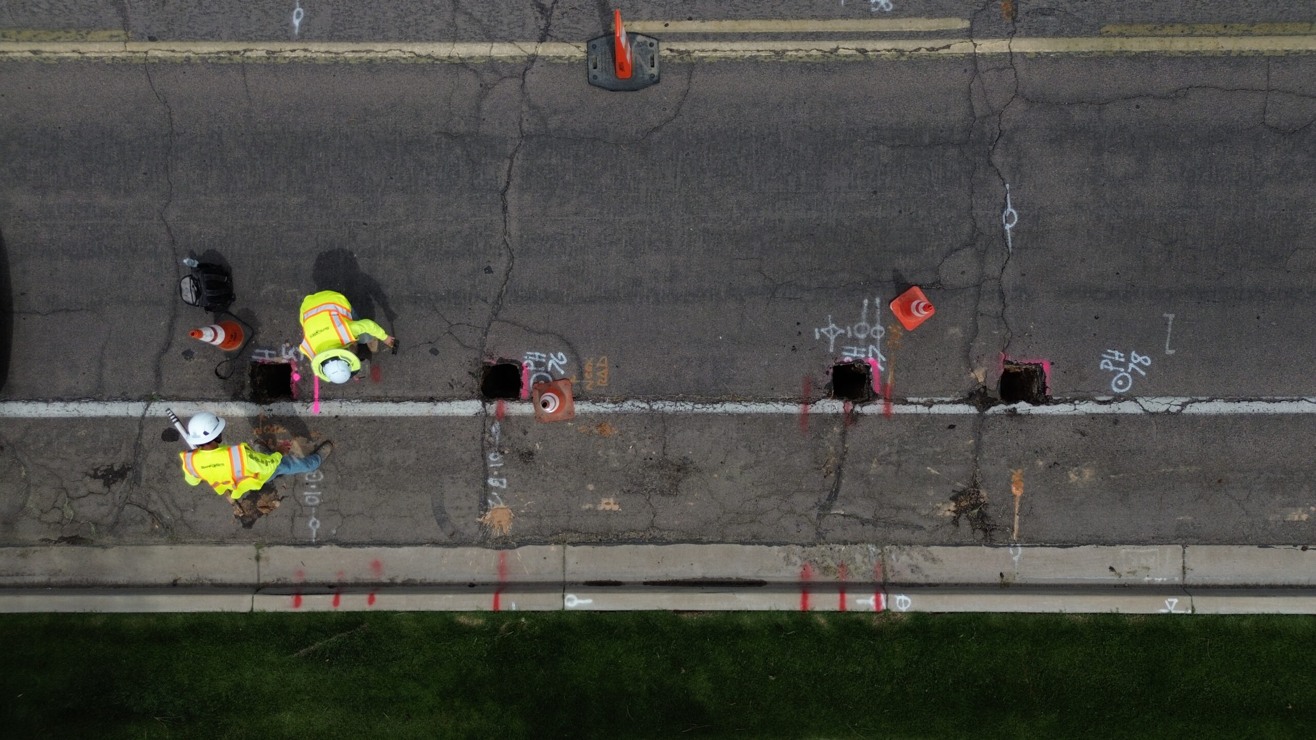 Aerial view of two construction workers in high-visibility vests and hard hats working beside several evenly spaced holes cut into an asphalt road, marked with spray paint and surrounded by traffic cones.