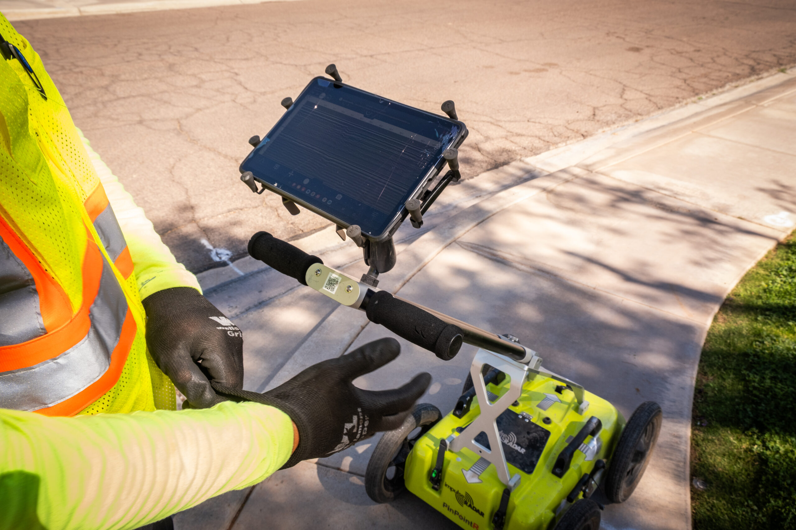 A close-up of a person operating a yellow ground-penetrating radar device with a mounted tablet, standing on a sidewalk.