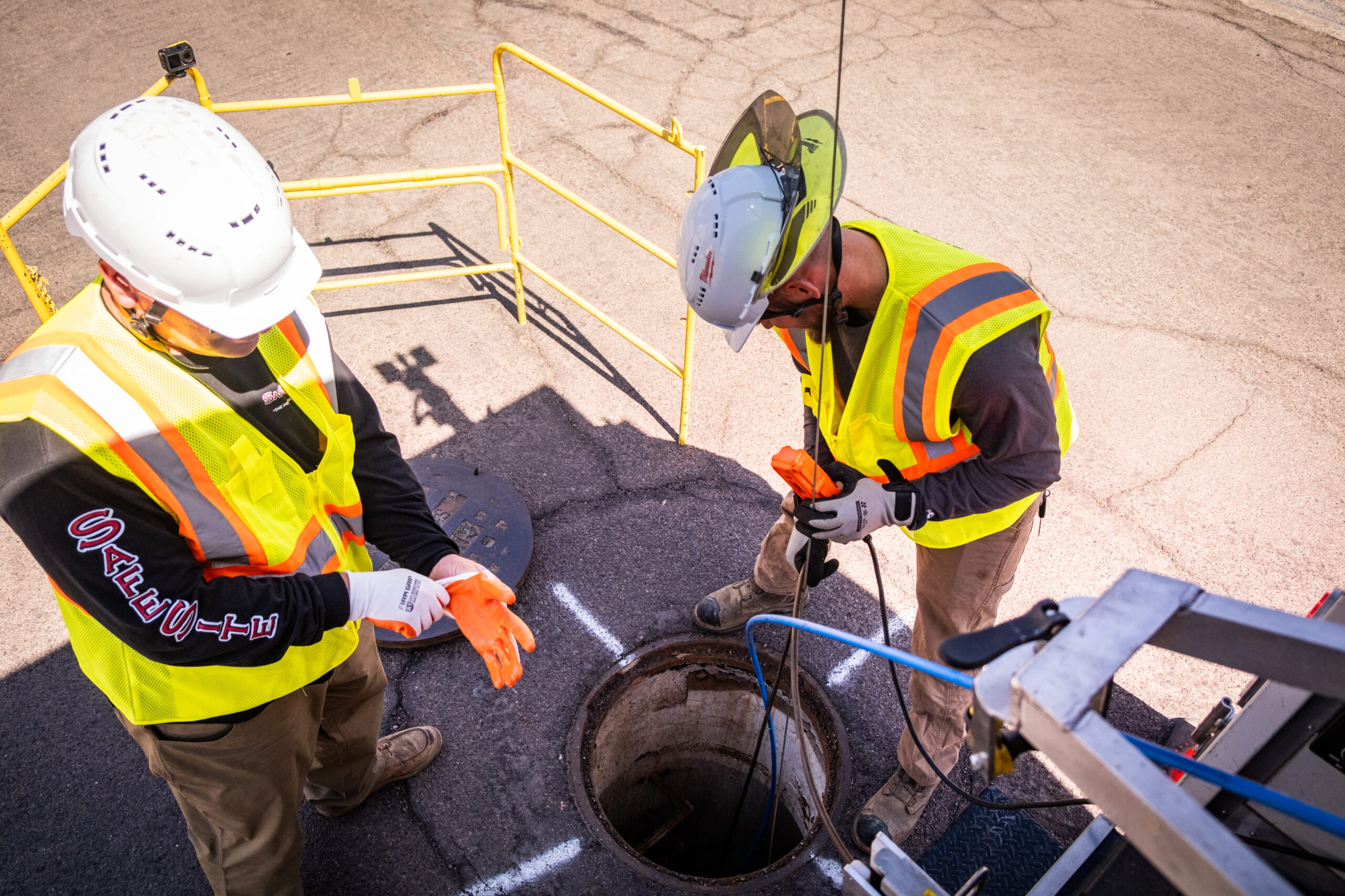 Two construction workers in high-visibility vests and hard hats stand next to an open manhole, one adjusting gloves and the other lowering equipment into the hole, with safety railings surrounding the work area.